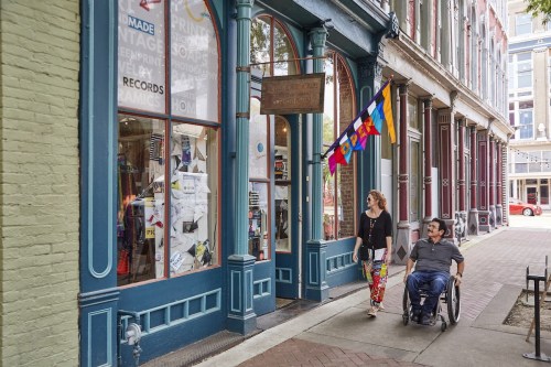 A woman and a man in a wheelchair walk past a colorful storefront with flags and displays in a vibrant urban setting.