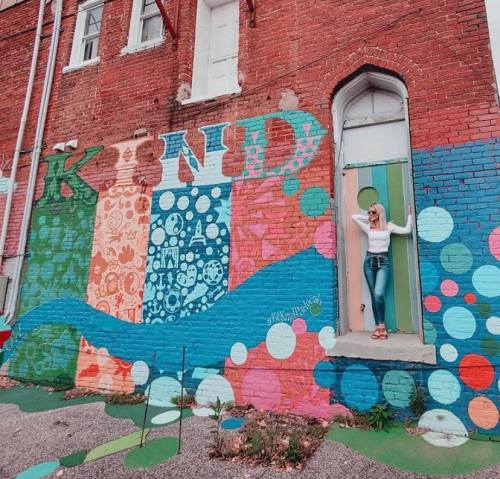 A person stands in a doorway beside a colorful mural that spells "KIND" with various patterns and designs.