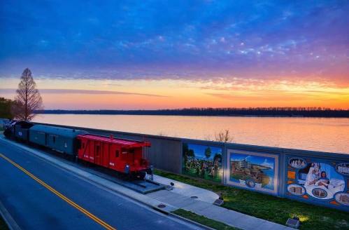 A red train caboose beside a river at sunset, with a mural on the wall and colorful clouds in the sky.
