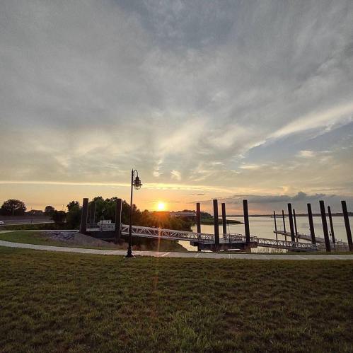 Sunset over a calm lake, with a dock and grassy area in the foreground, and clouds in the sky.