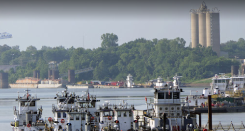 Tugboats and barges on a river with green hills and silos in the background on a clear day.