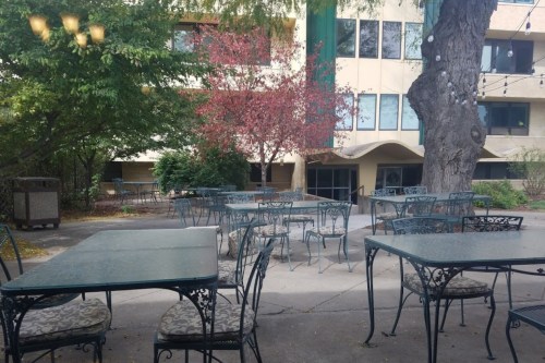 Outdoor seating area with metal tables and chairs surrounded by trees and a building in the background.