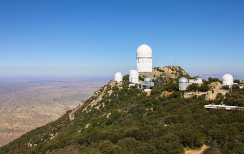 A mountain observatory with large domes, surrounded by green hills and a vast desert landscape under a clear blue sky.