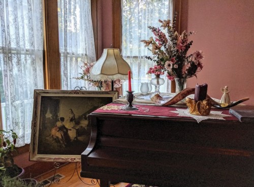 A cozy room with a piano, vintage lamp, flowers, a painting, and decorative items on a table by the window.
