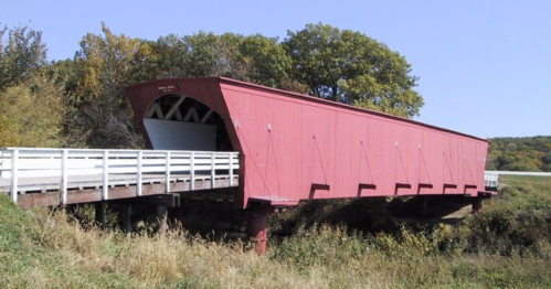 A red covered bridge spans a grassy area, surrounded by trees under a clear blue sky.