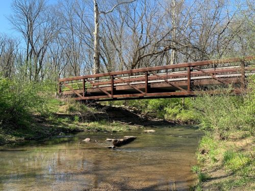 A wooden bridge spans a clear creek, surrounded by trees and greenery under a bright blue sky.