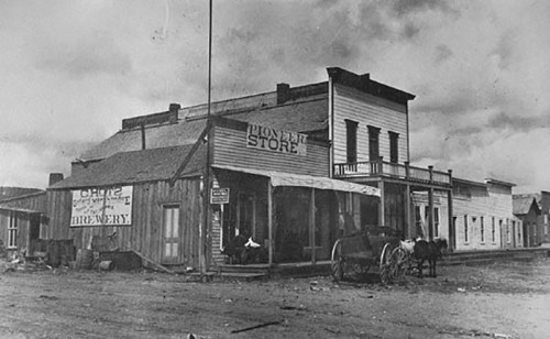 Historic black-and-white photo of a pioneer store and brewery in a small town, with a horse-drawn carriage outside.