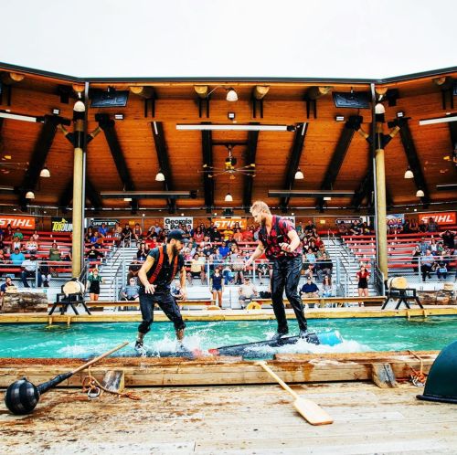 Two men in life jackets perform on a water stage, surrounded by an audience in a wooden amphitheater.