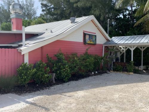 A pink building with a metal roof, surrounded by greenery and gravel, under a sunny sky.