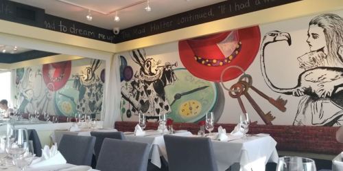 Colorful mural featuring whimsical characters and objects, set in a stylish restaurant with elegant table settings.