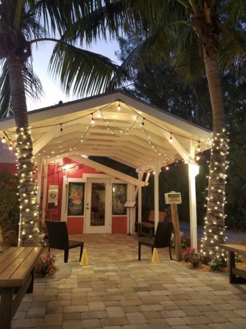 A charming entrance with a white canopy, palm trees, and string lights, surrounded by colorful flowers and paver stones.