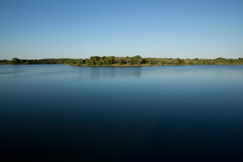A calm lake reflecting a clear blue sky, with a small island of greenery in the distance.