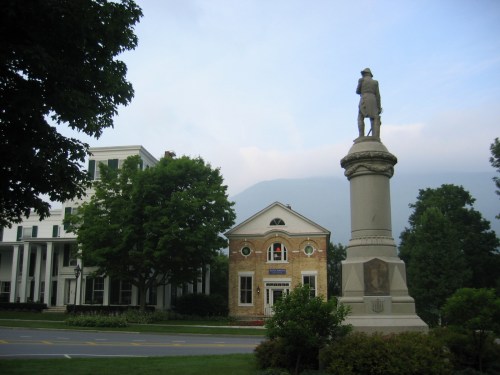 A statue stands in front of a historic building, surrounded by trees and mountains in the background.