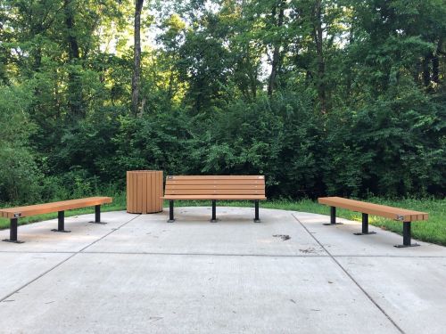 A seating area with wooden benches and a trash can, surrounded by lush green trees in a park setting.