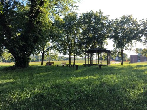 A peaceful park scene with green grass, trees, benches, and a gazebo under a clear blue sky.