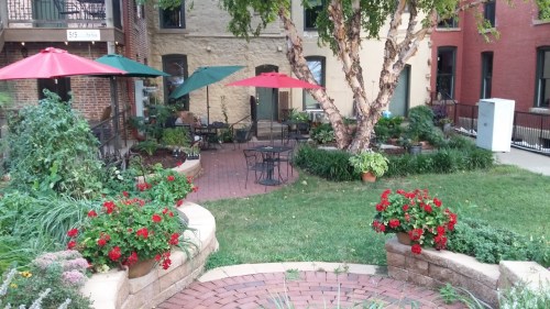 A cozy courtyard with red and green umbrellas, surrounded by lush plants and flowers, featuring a brick patio and seating.