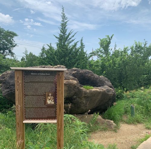 A large rock formation beside a sign detailing the history of Indian Rock, surrounded by greenery and a pathway.