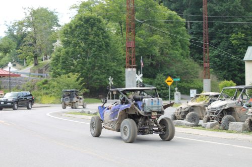 A muddy off-road vehicle drives along a road with trees and other vehicles in the background.