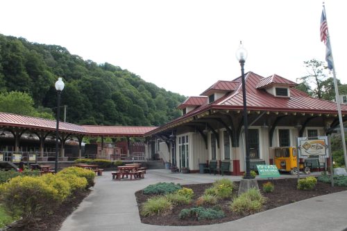 Historic building with a red metal roof, surrounded by greenery and picnic tables, set against a wooded hillside.