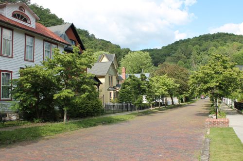 A peaceful street lined with trees and charming houses, set against a backdrop of green hills under a blue sky.