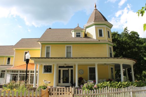 A charming yellow Victorian house with a turret, surrounded by greenery and a white picket fence.
