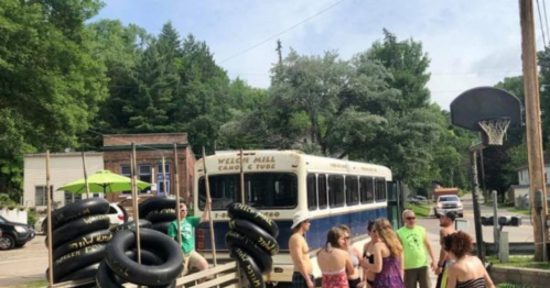 A group of people gathers near a bus and inflatable tubes in a sunny outdoor setting with trees in the background.