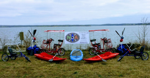 A display of various watercraft, including kayaks and pedal boats, by a lake with a promotional banner in the background.