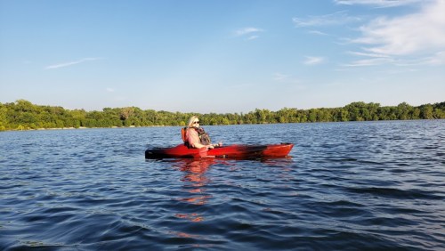 A person in a red kayak paddles on a calm lake surrounded by green trees under a clear blue sky.