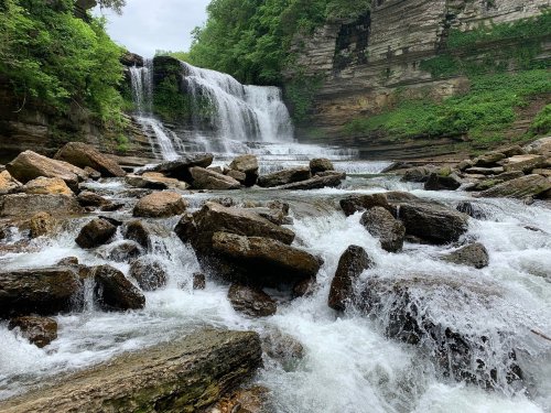 A serene waterfall cascades over rocky cliffs, surrounded by lush greenery and flowing water over stones in the foreground.