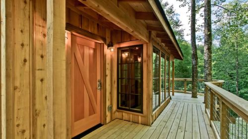 A wooden cabin entrance with a large door and windows, surrounded by trees and a wooden deck.