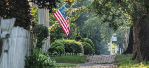 A peaceful street scene with a U.S. flag hanging, lined with greenery and a cobblestone path.