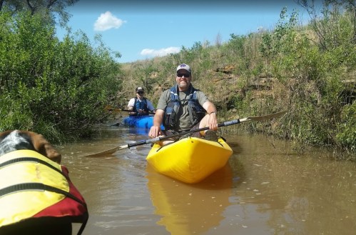 Two people kayaking in a narrow waterway surrounded by greenery, with a dog in a life jacket in the foreground.