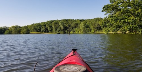 A red kayak on calm water, facing a lush green shoreline under a clear blue sky.
