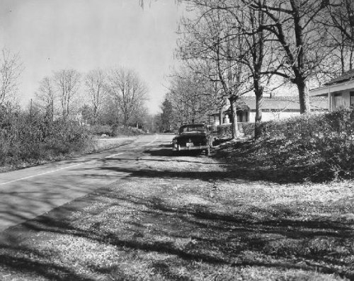 A black-and-white photo of a quiet road lined with bare trees and a parked car beside a house.