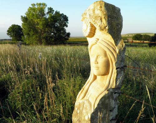 A carved stone sculpture of a woman stands in a grassy field, with a tree and blue sky in the background.