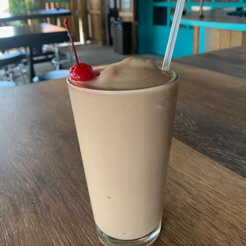 A creamy beverage in a tall glass, topped with a cherry, sits on a wooden table in a casual setting.