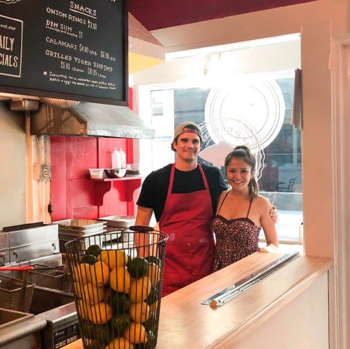 A man in an apron and a woman in a dress stand smiling in a bright, casual eatery with a basket of lemons.