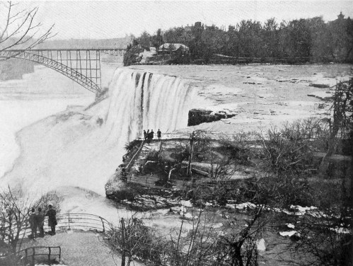 Historic black-and-white photo of Niagara Falls, featuring a bridge and people near the water's edge.