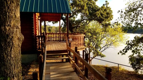A wooden cabin with a blue roof overlooks a serene lake, surrounded by trees and a wooden walkway.