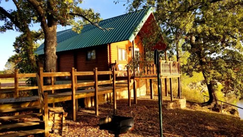 A cozy wooden cabin with a green metal roof, surrounded by trees and a wooden deck overlooking a serene landscape.