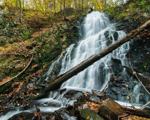 A serene waterfall cascades over rocky terrain, surrounded by autumn foliage and fallen logs.