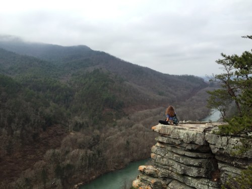 A person sits on a rocky ledge overlooking a river and forested mountains under a cloudy sky.