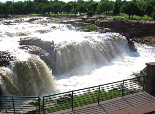 A scenic view of a waterfall cascading over rocky cliffs, surrounded by lush greenery and a park area.