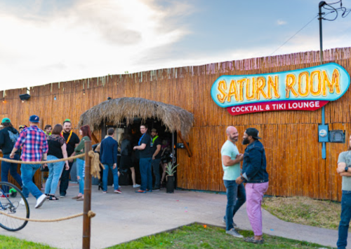 A lively outdoor scene at the Saturn Room cocktail and tiki lounge, with people socializing near a bamboo-covered entrance.