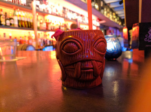 A tiki mug with a face design sits on a bar counter, surrounded by colorful lights and bottles in the background.