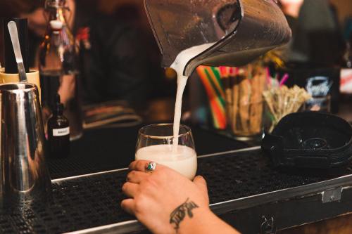 A bartender pours a creamy drink from a shaker into a glass at a lively bar.