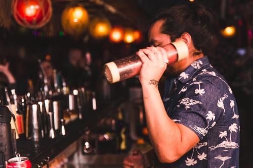 A bartender in a floral shirt shakes a cocktail at a vibrant bar with colorful lights and various bottles in the background.