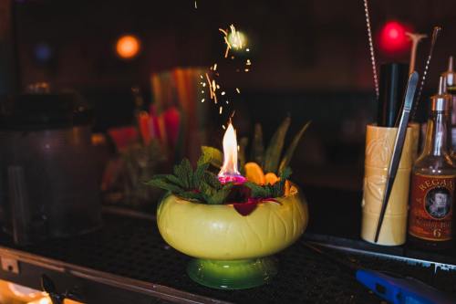 A vibrant cocktail garnished with fresh herbs and flowers, featuring a small flame and sparks, set on a bar counter.