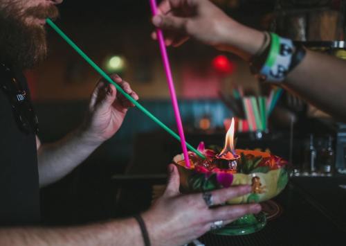 A person holds a colorful drink with a flaming garnish, while another adds a straw. Dimly lit bar atmosphere.