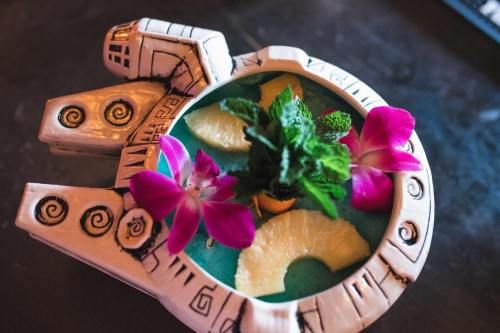 A colorful cocktail served in a decorative hand-shaped glass, garnished with pineapple, mint, and purple flowers.